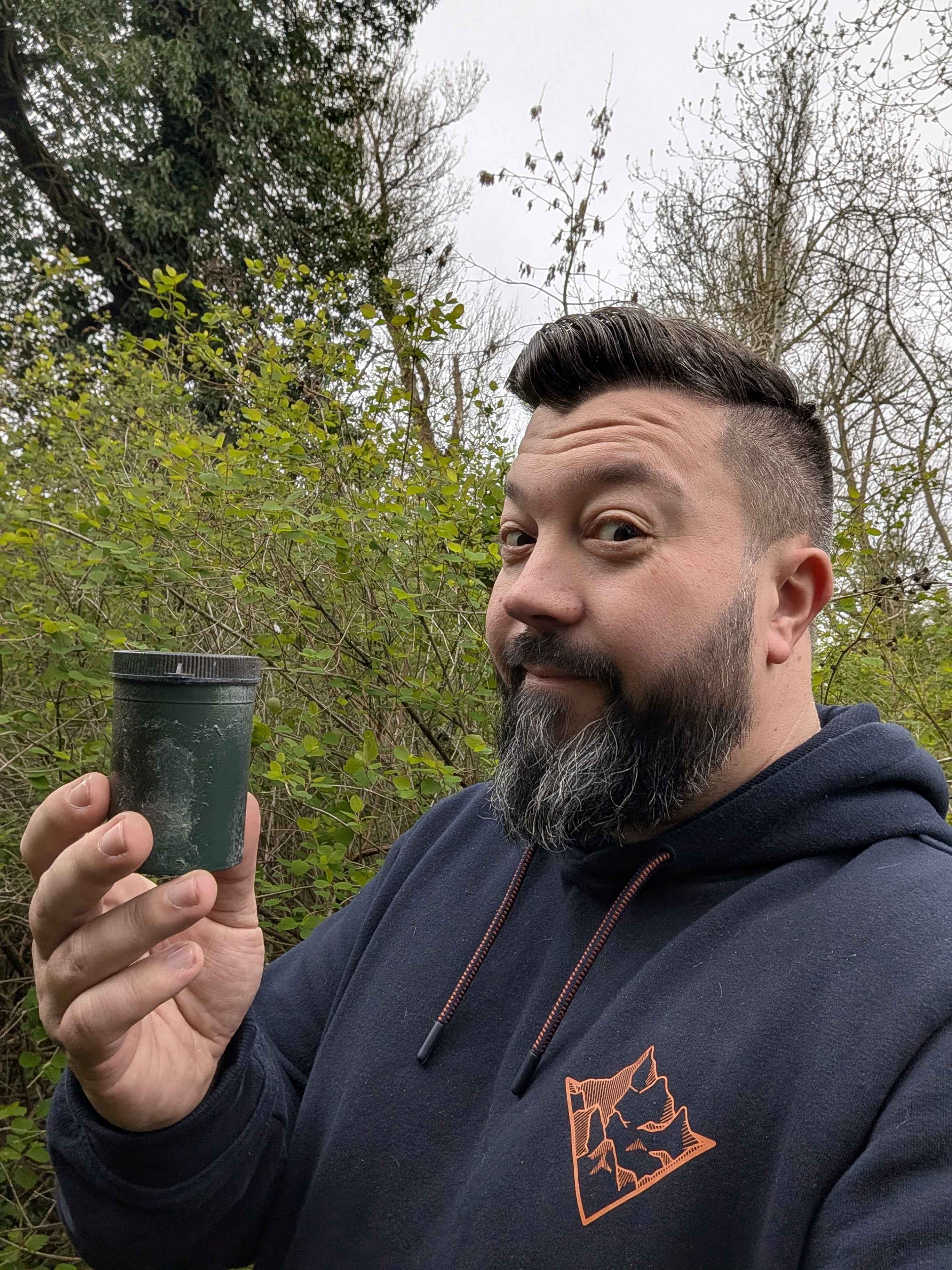 Gaz standing in front of hedge with small green geocache container